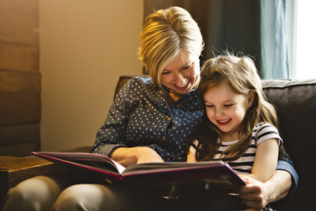 A mother teaching daughter holding book sitting on sofa